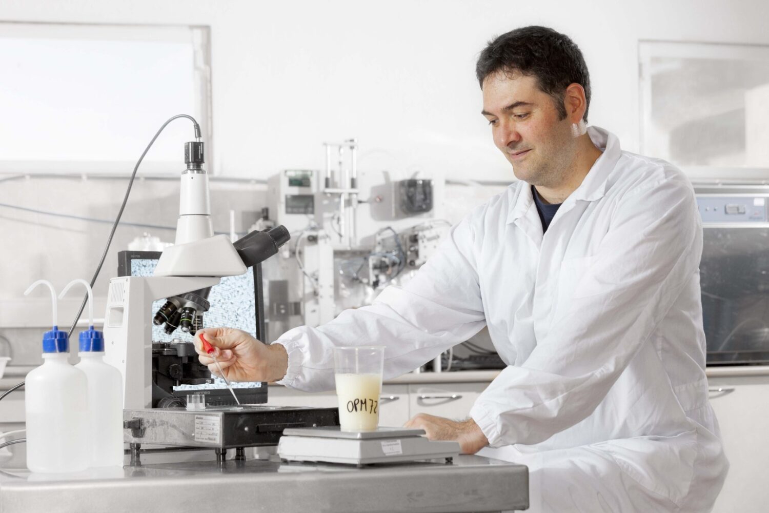Een wetenschapper in een witte labjas onderzoekt een monster onder een microscoop in een laboratorium, met labapparatuur en een bekerglas met het label "ORM" zichtbaar op de tafel.