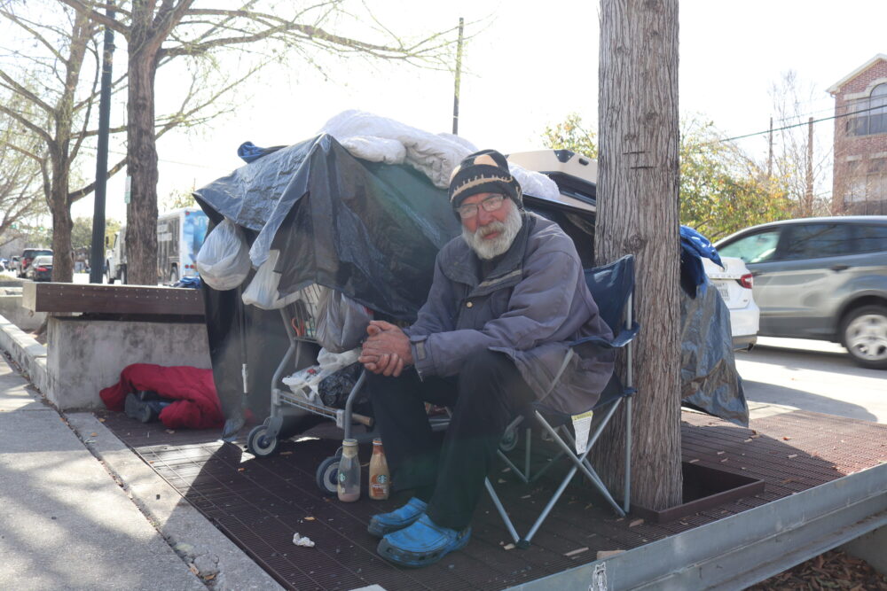 Dave, a homeless man, sits along Bagby Street next to another sleeping homeless man in Midtown in March, 2025. Midtown is one of the 12 areas in Houston where such behavior is illegal during daylight hours. (Dominic Anthony Walsh/HPM)