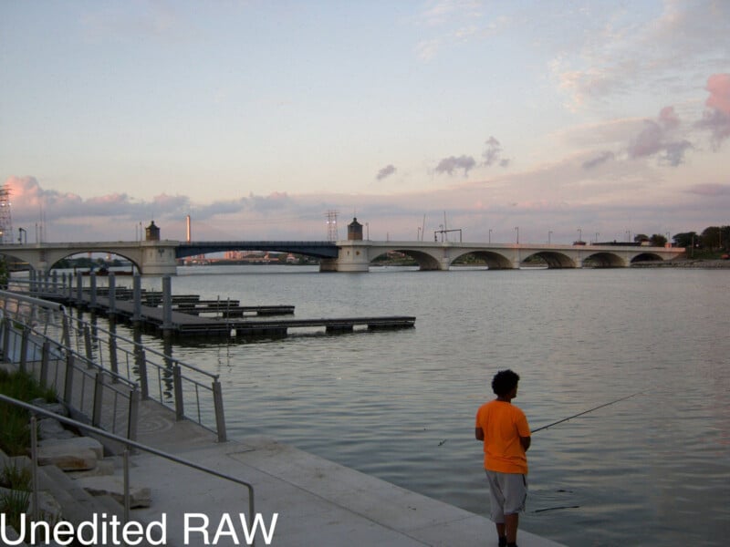 A person in an orange shirt fishes by the waterfront near a dock, with a large arched bridge in the background under a partly cloudy sky. The words "Unedited RAW" appear in the lower left corner.
