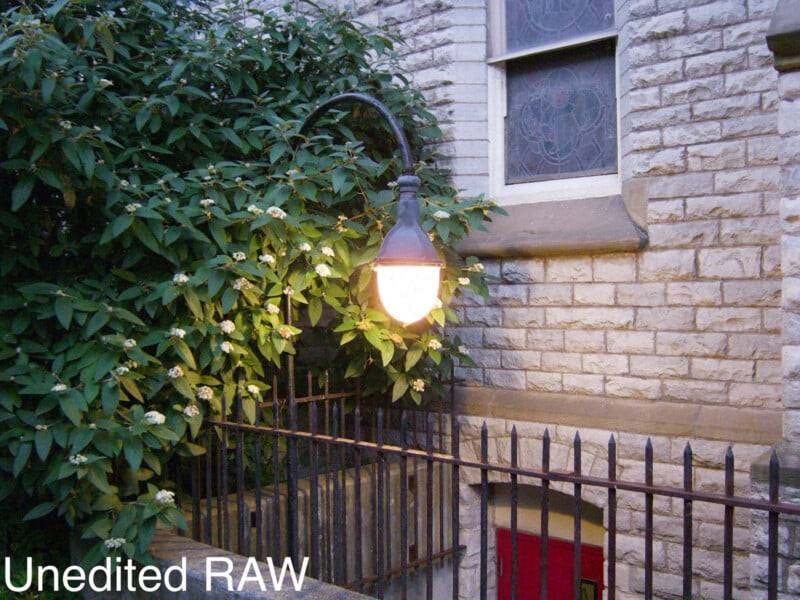 An old-fashioned streetlamp glows beside a leafy green bush with small white flowers, in front of a stone building with a stained glass window and a red door. A black iron fence runs along the foreground. Text reads "Unedited RAW.