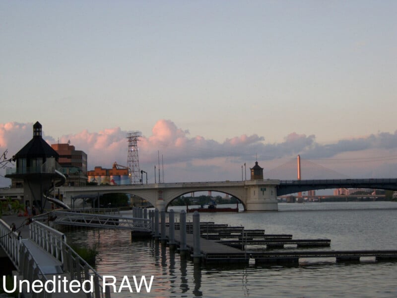 A calm river with floating docks in the foreground, a bridge and industrial buildings in the background, and partly cloudy skies at sunset. Text reads "Unedited RAW" in the bottom left corner.