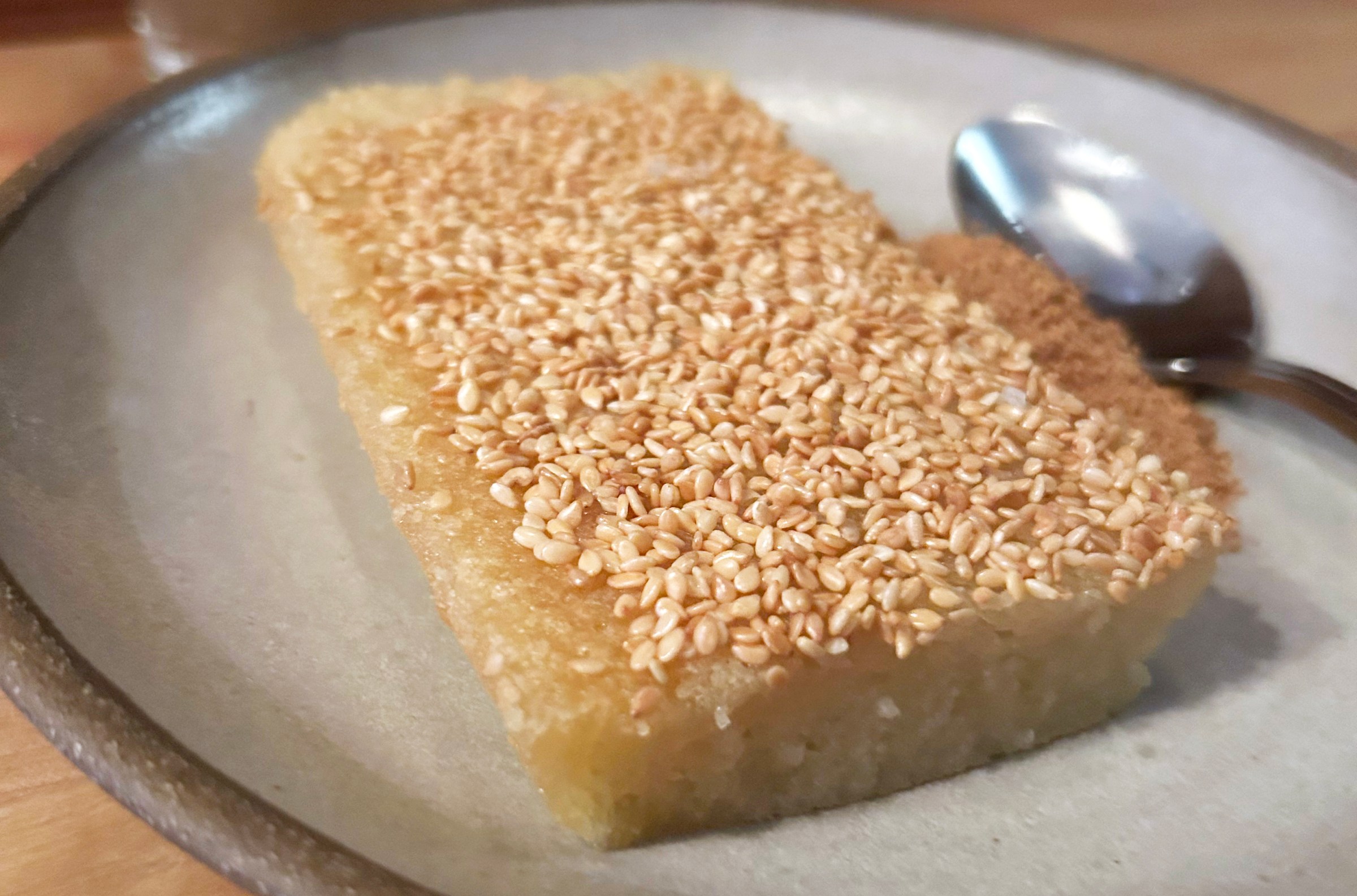 A butter-yellow mochi cake with sesame seeds on top and a pile of brown dust behind it on a plate.