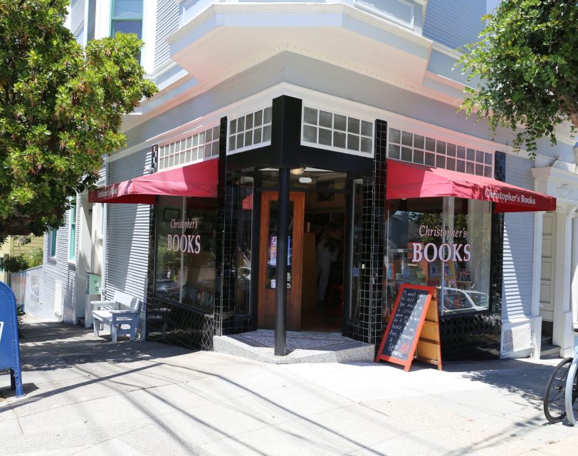 Street view of a corner bookstore with a red awning labeled "Books," large windows, a chalkboard sign outside, and bicycles parked nearby.