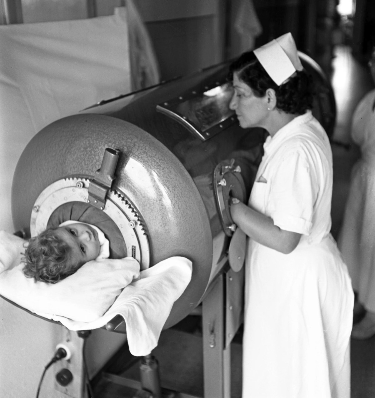 A nurse monitors a young child lying inside an iron lung machine, used for respiratory support, in a hospital setting.