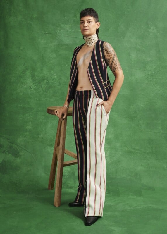 A person with short hair stands confidently beside a tall wooden stool, wearing a sleeveless, open pinstripe vest and matching wide-leg pants. Their chest is partially exposed, displaying a top surgery scar and tattoos. The background is green.