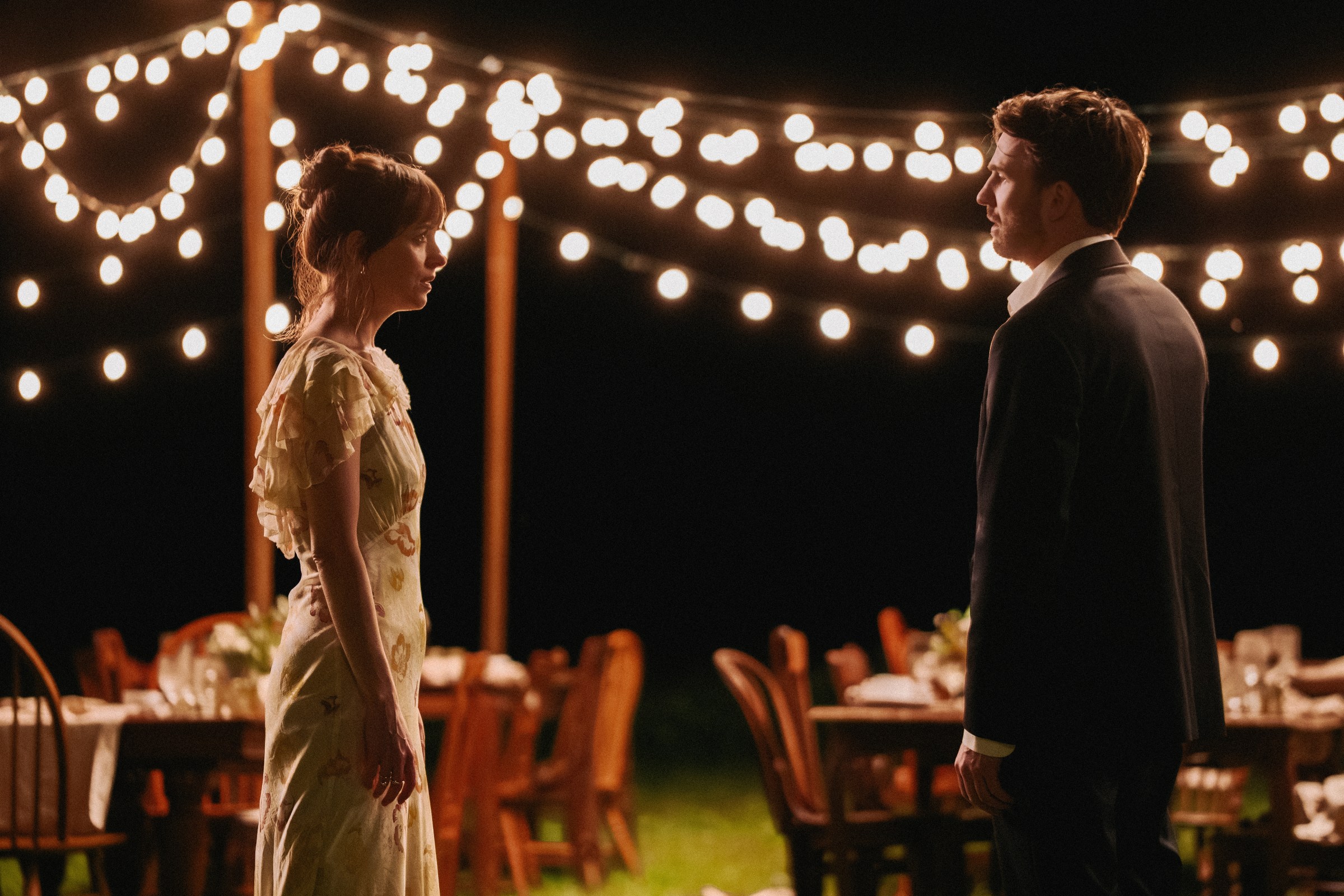 Two people outside in front of tables and strung lights at night.