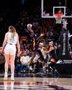 The image depicts a basketball game in progress with players in motion near the hoop. A player in a black uniform with colorful accents is in mid-air, holding the basketball with an outstretched arm, poised to make a shot. 
