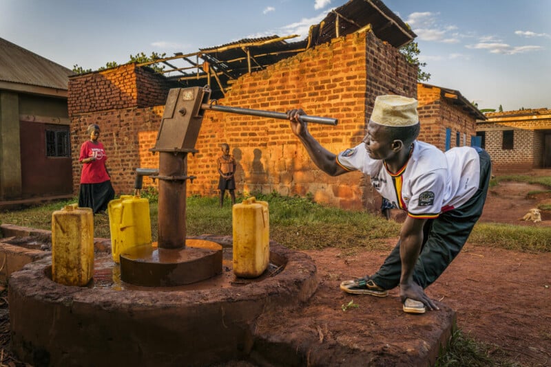 A man in a white shirt and hat pumps water into yellow jerry cans at a hand water pump in a rural village, with children and brick buildings in the background under a blue sky.