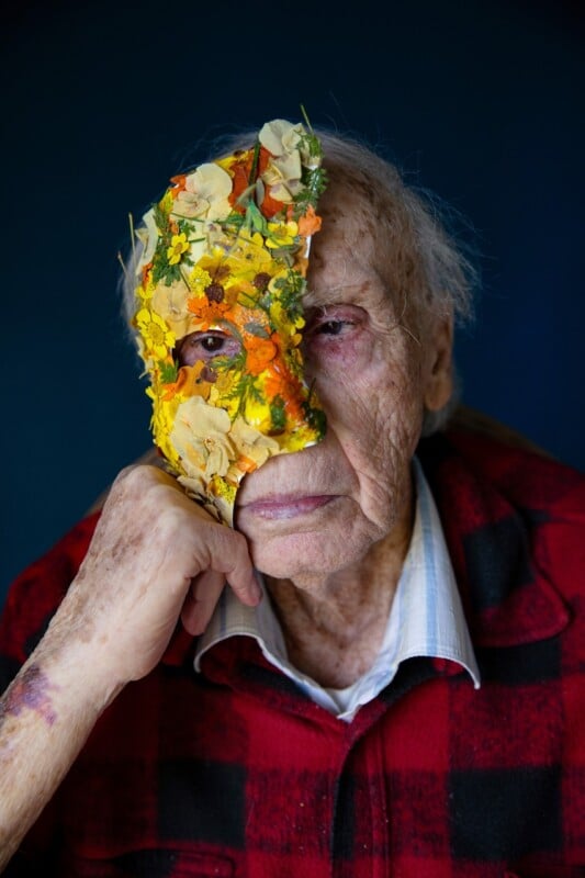 An elderly person with gray hair wears a vibrant half-mask made of flowers and leaves on the left side of their face, gazing thoughtfully at the camera. They wear a red and black plaid shirt.