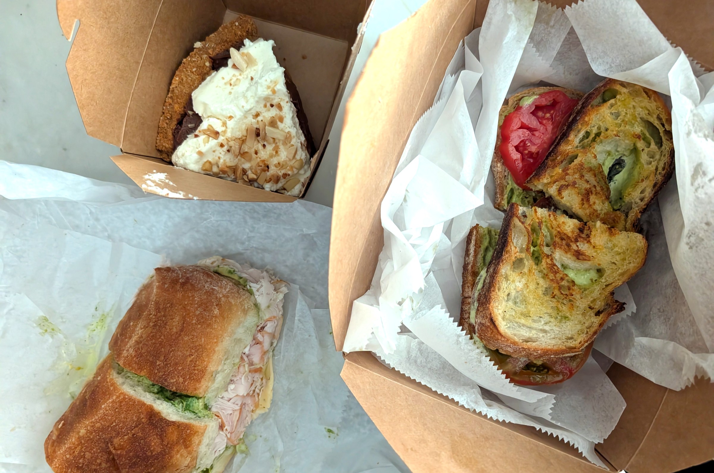 A sandwich on a wax paper next to a sandwich in a cardboard container next to a slice of cream-topped pie in another cardboard box.
