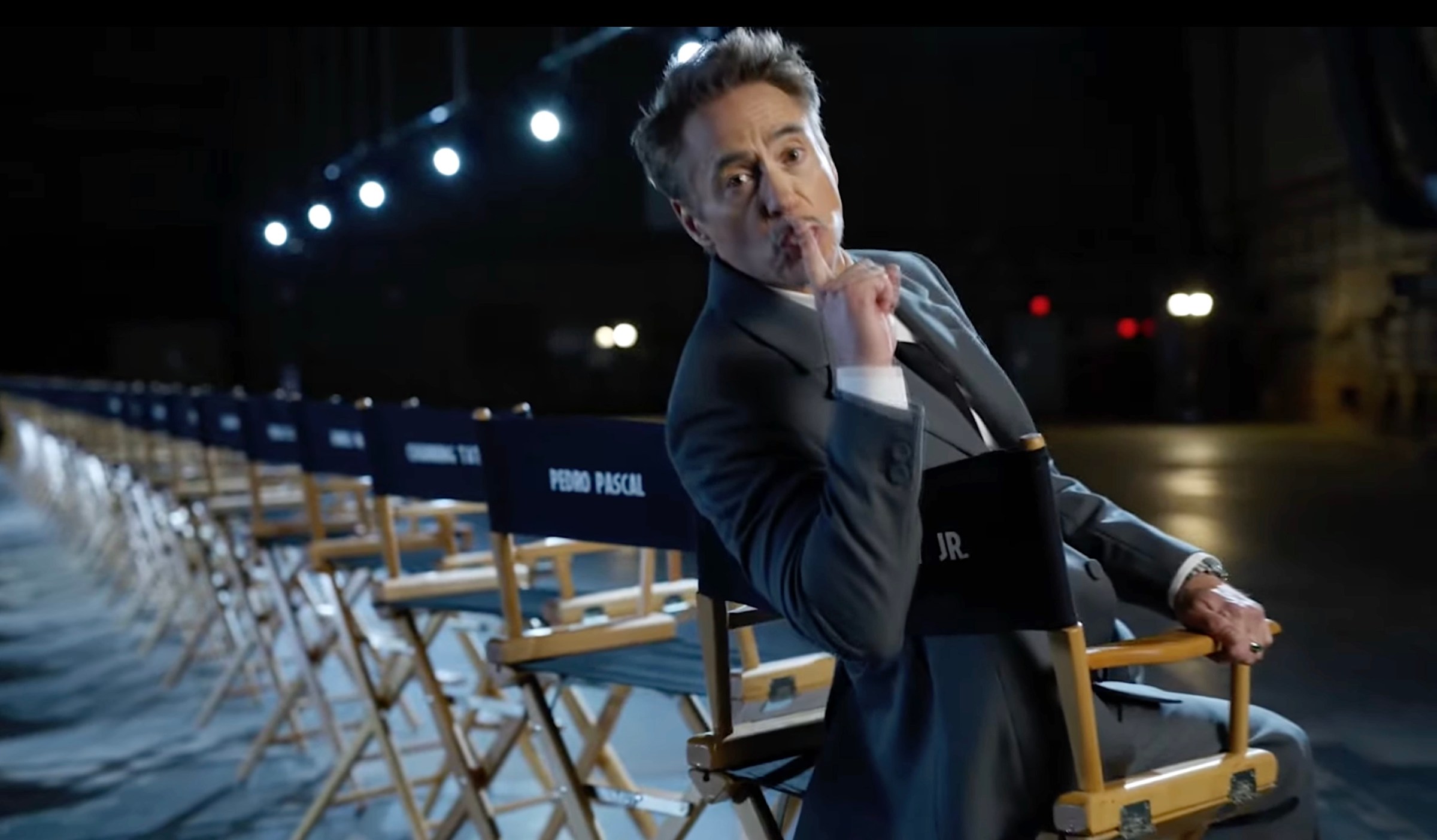 Robert Downey Jr. sits in a director’s chair in a long line of similar chairs on a dark soundstage, pressing a finger to his lips in a “shush” gesture, in Marvel’s livestream announcement of the Avengers: Doomsday cast