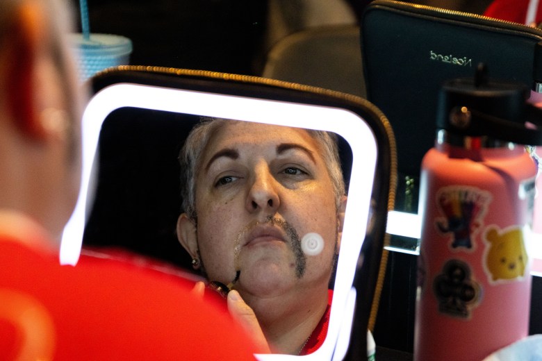 A face in a mirror with water bottle and clutter around shows a middle aged person with half a mustache on. 