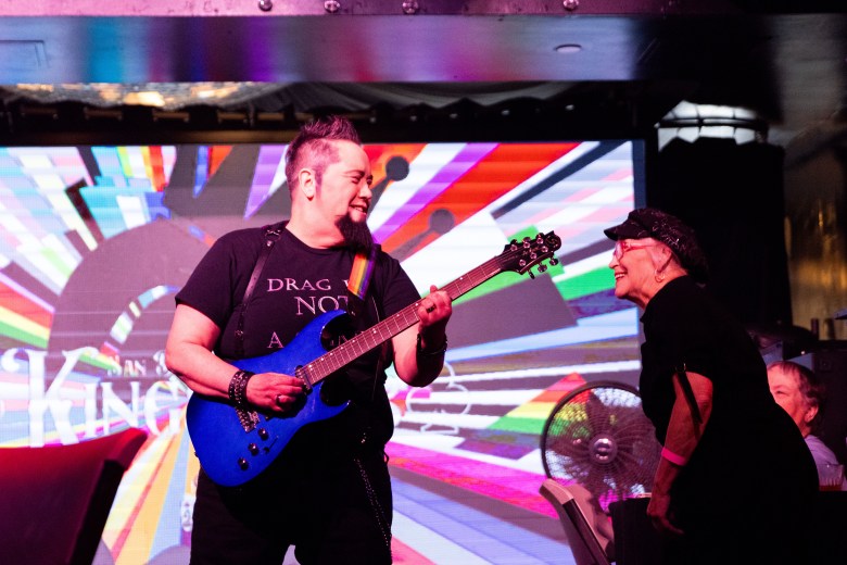 A man in a goatee playing an electric guitar looks at a standing elderly woman wearing black. 