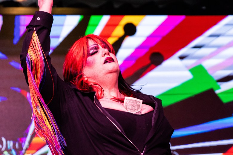 A woman with a red wig closes her eyes and lifts up her arms. Bills are peeking out between her breasts. 