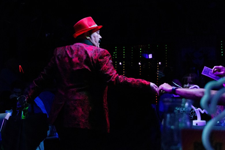 A person in drag wearing a red suit and red hat reaches over to an audience member holding a dollar. 