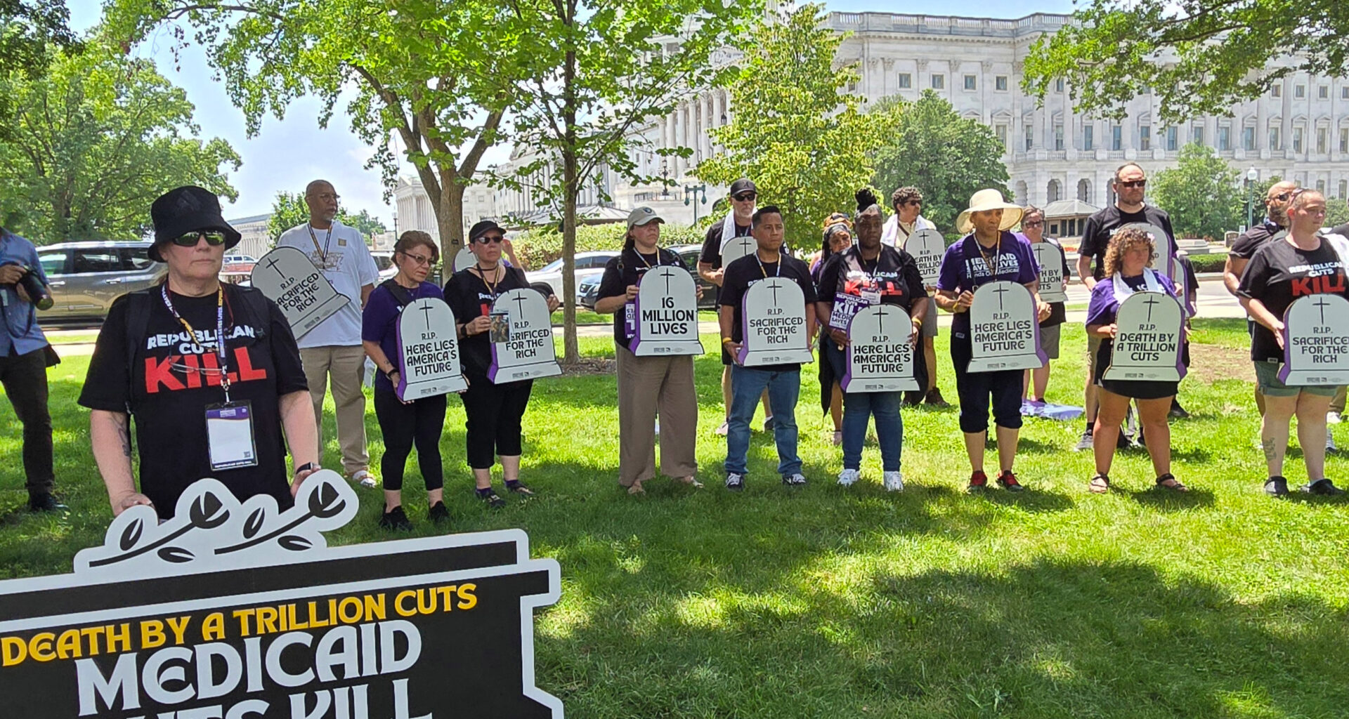A group of protesters stand holding signs. One large sign reads, "Death by a trillion cuts: Medicaid cuts kill." Others hold signs shaped like tombstones that read, "Here lies America's future," and "R.I.P. Sacrificed for the rich."