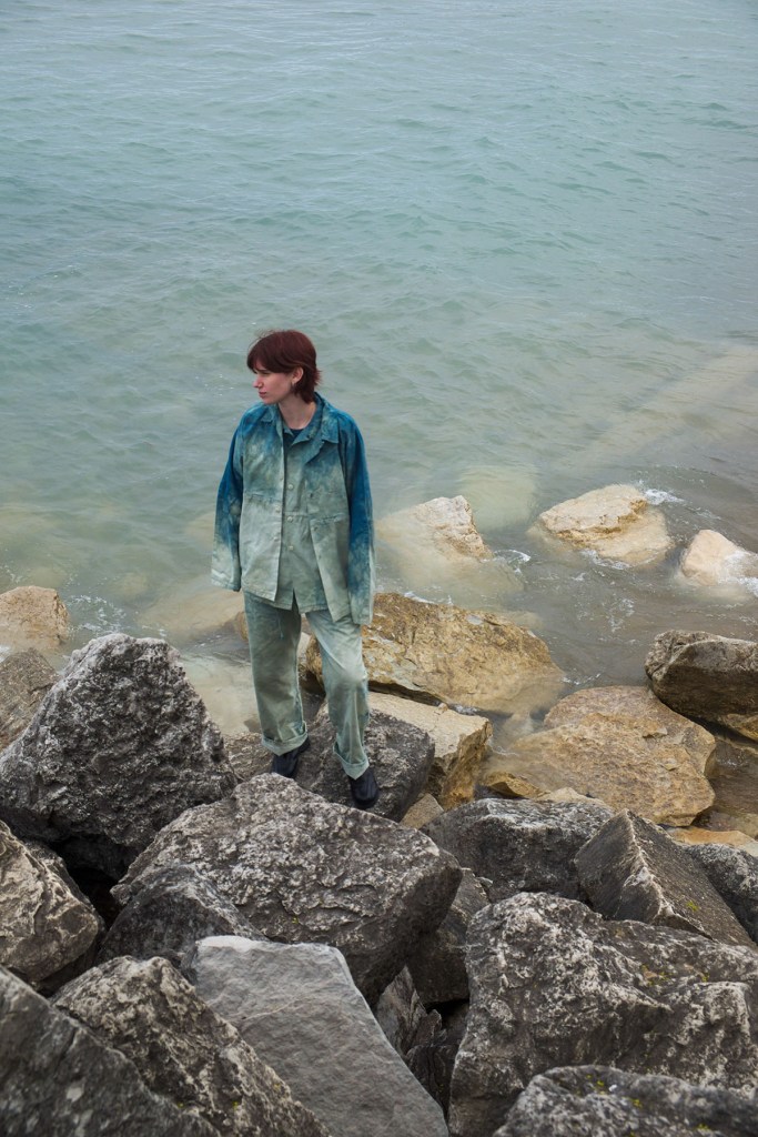 a model stands on rocks next to a body of water while wearing a hip-length denim jacket and matching denim jeans
