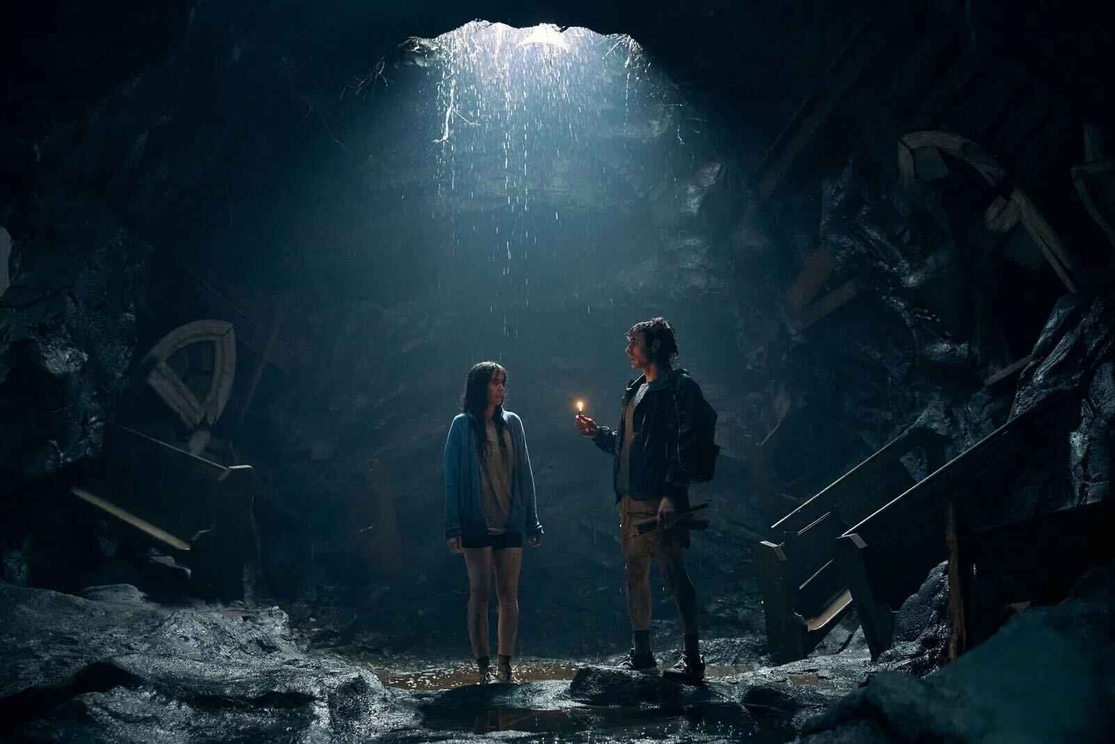 Alison Brie and Dave Franco in Together, standing in a dark, damp cave as rain descends on them from a hole in the cavern’s ceiling above them