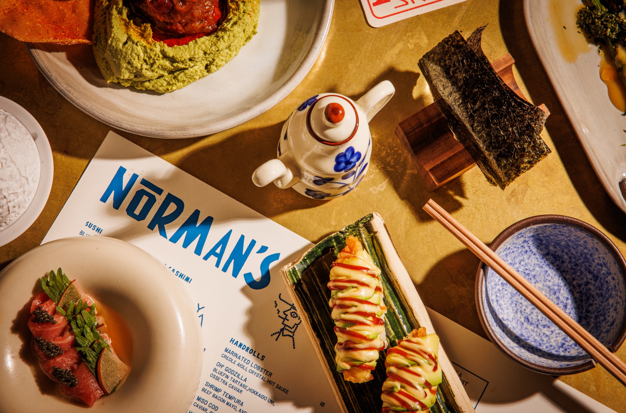 A dish with nigiri, a container that looks like a mini-tea pot, chopsticks, and edamame hummus sit on a table.