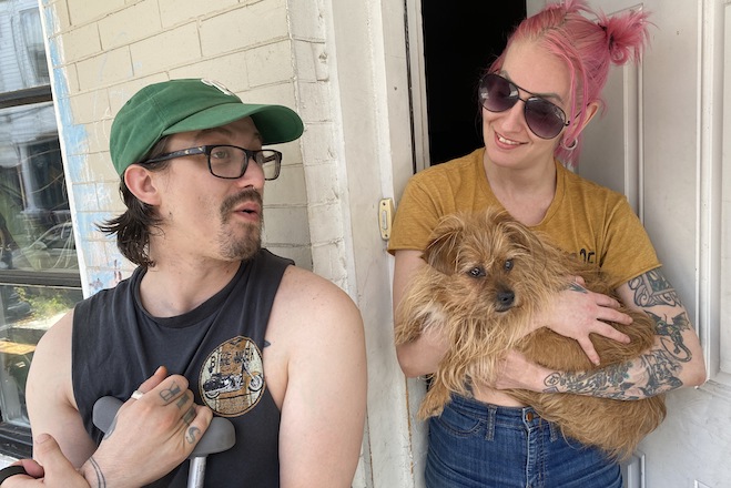 Dennis Petrakis and Sara Crosby, a white man and woman; he's in glasses with a green Phillies cap, finger tattoos, a sleeveless shirt, holding a crutch; she has pink hair, large purple sunglasses, a tattooed arm holding a small Yorkie, with dog Eevee discussing the mass shooting on their block at their front door.