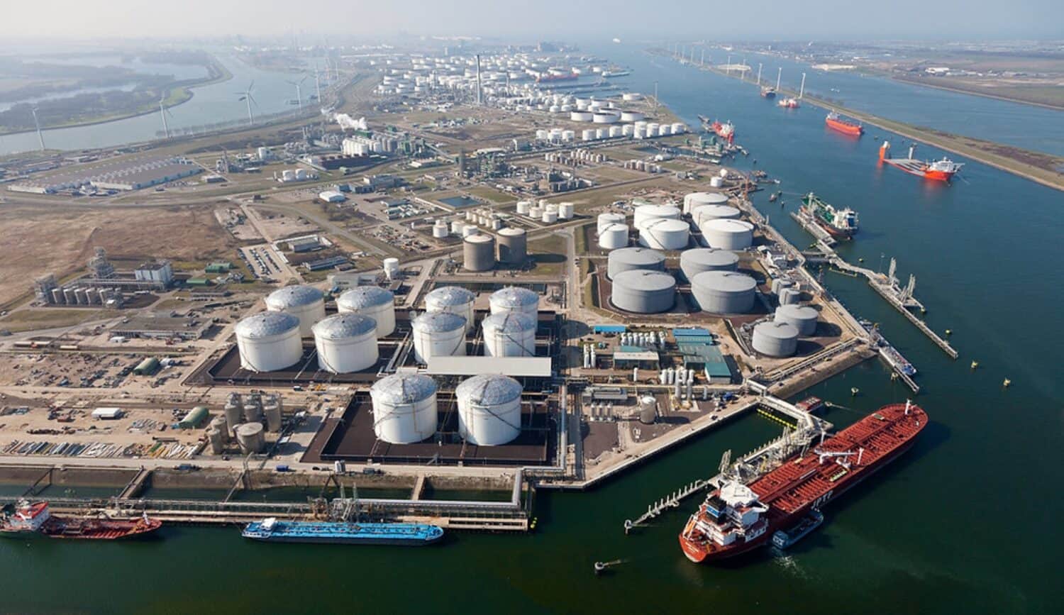 Luchtfoto van een grote industriële haven met olieopslagtanks, aangemeerde vrachtschepen, pijpleidingen en waterkanalen.