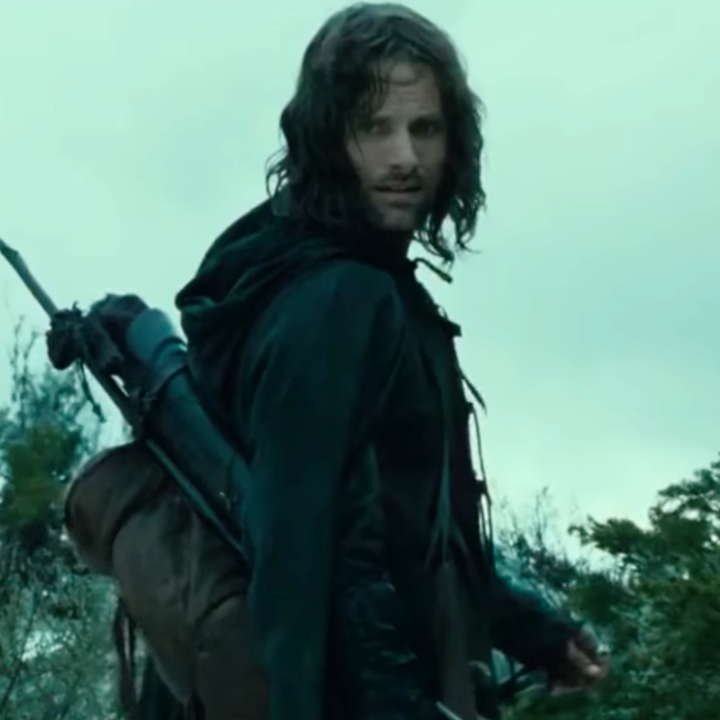 A person with long hair and a beard in an outdoor setting, carrying a quiver of arrows and looking back, dressed in medieval-style clothing