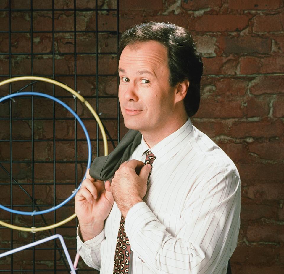Person in a pinstripe shirt and patterned tie, playfully adjusting their jacket. Brick wall with neon art in the background