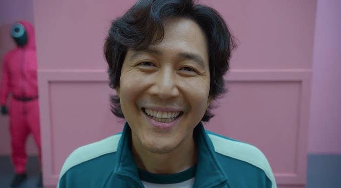 Smiling man in a teal tracksuit poses in front of a pink wall, with a person dressed in a pink jumpsuit and mask in the background