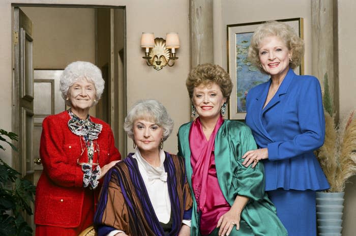 Four women pose together, each dressed in elegant 1980s-style clothing, in a living room setting