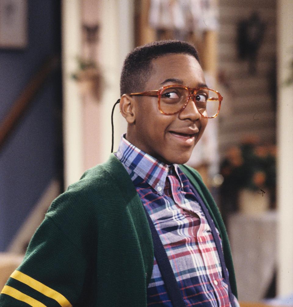 Person wearing a plaid shirt, suspenders, and a green cardigan with yellow stripes, looks cheerful on a TV set