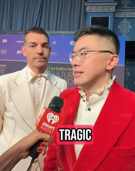 Two individuals on a press event stage. The person in front wears a red suit with a ruffled shirt, speaking into an iHeartRadio microphone. The word "TRAGIC" appears