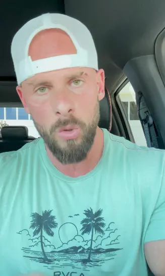 Man with cap and beard talks in a car, wearing a tropical-themed T-shirt