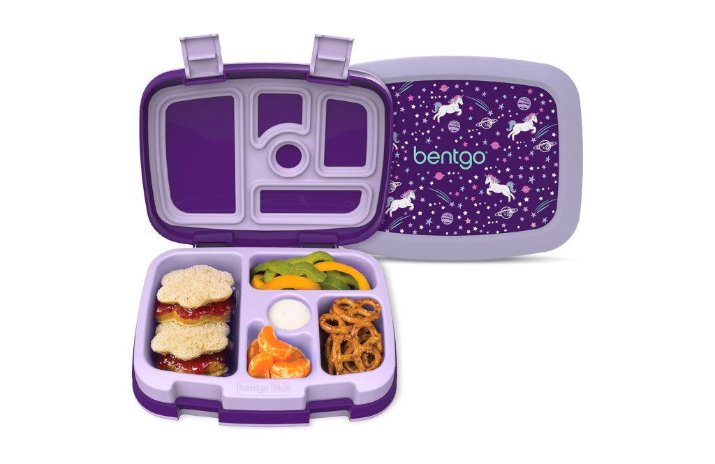 Purple bento box with compartments containing peanut butter sandwiches, bell peppers, mandarin oranges, pretzels, and dip; the lid features a unicorn and star design.