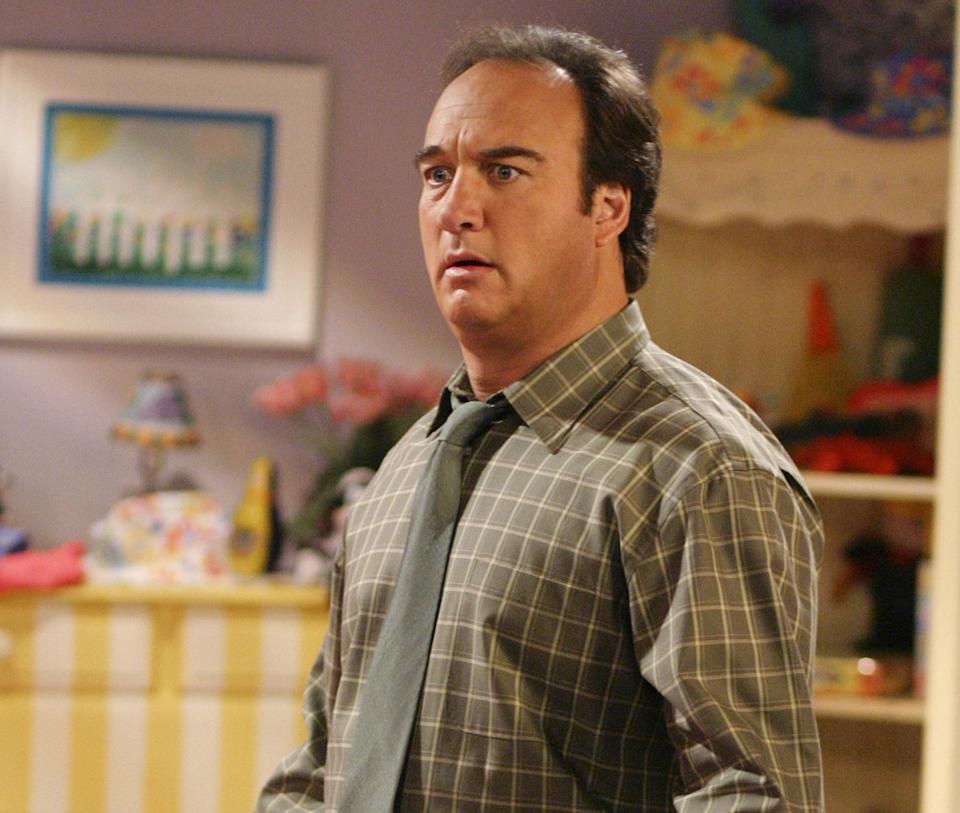 A person in a plaid shirt and tie looks surprised while holding a remote, in a home setting with decorative shelves in the background