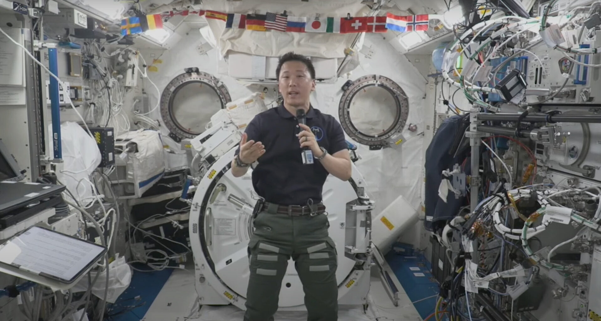 an astronaut wearing a dark blue polo shirt speaks into a microphone inside a cluttered space station module