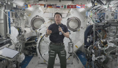 an astronaut wearing a dark blue polo shirt speaks into a microphone inside a cluttered space station module