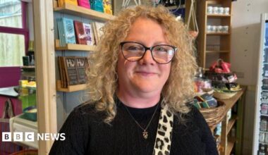 Cherylee Barker smiles at the camera. She is stood in a cafe which shelves and chocolate bars in the background,