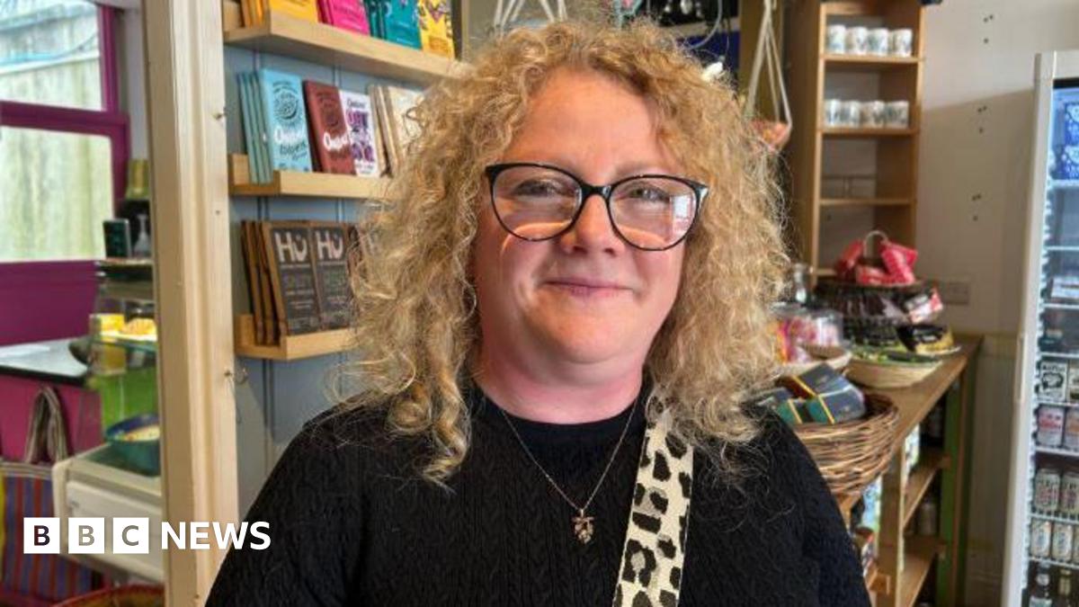 Cherylee Barker smiles at the camera. She is stood in a cafe which shelves and chocolate bars in the background,