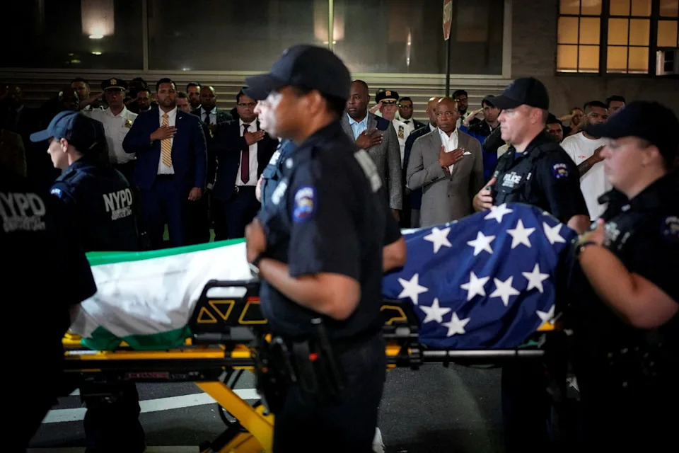 New York Mayor Eric Adams stands in a crowd of people as they watch the flag-draped body of Officer Islam wheeled by.
