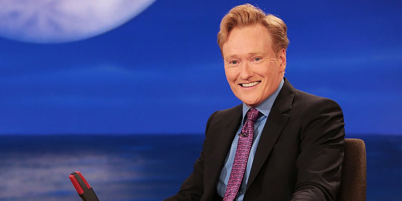 Conan O'Brien smiling behind his desk in Late Night with Conan O'Brien.