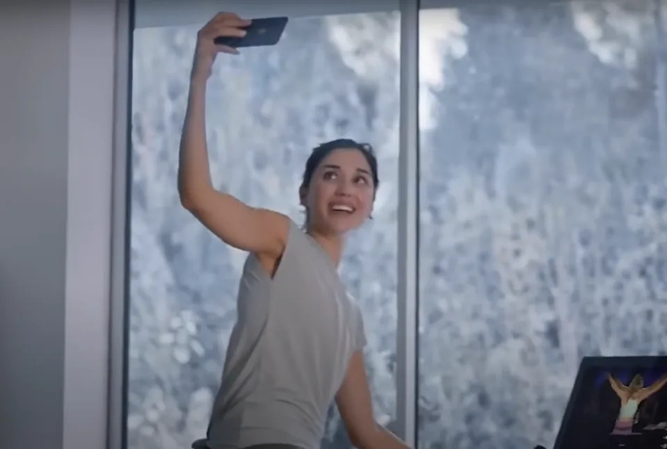 Person indoors smiling while taking a selfie, standing near a laptop, with a snowy landscape visible through a window