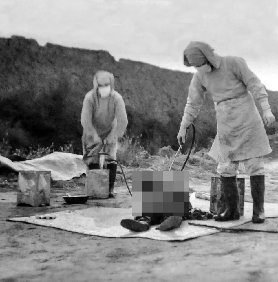Two people in protective gear perform a procedure on a child sitting on a tarp outdoors. The setting appears barren and remote