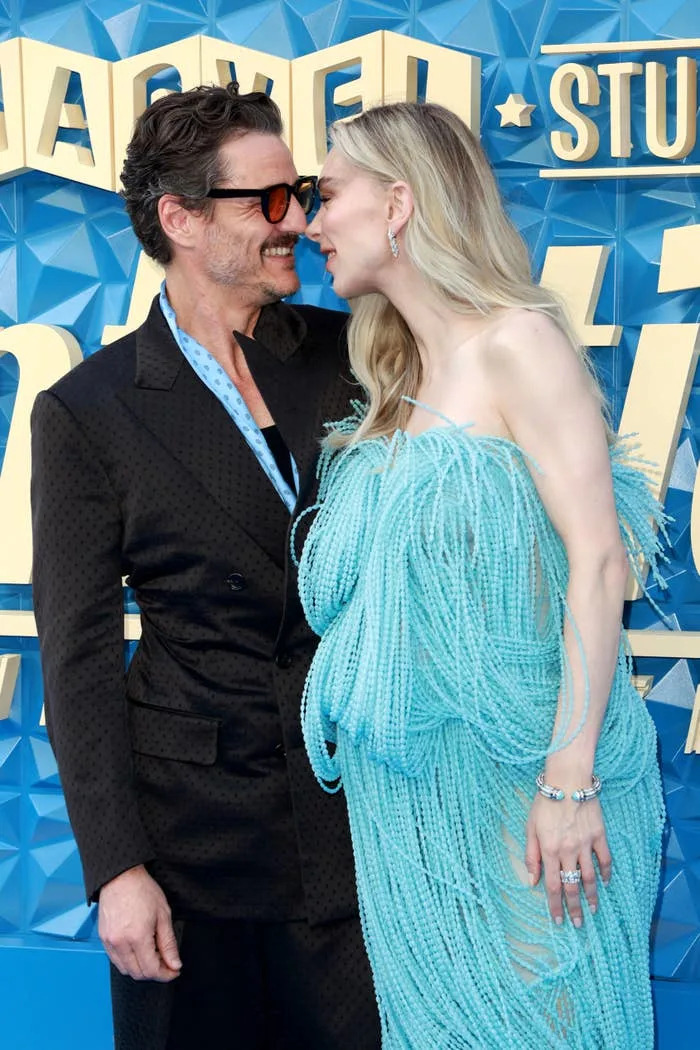 Two celebrities on a red carpet, one wearing a dark suit and glasses, the other in a flowing, textured dress, appear to be sharing a light moment