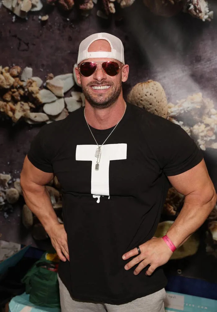 Man wearing a T-shirt, sunglasses, and a baseball cap, posing with hands on hips. Background features a textured design
