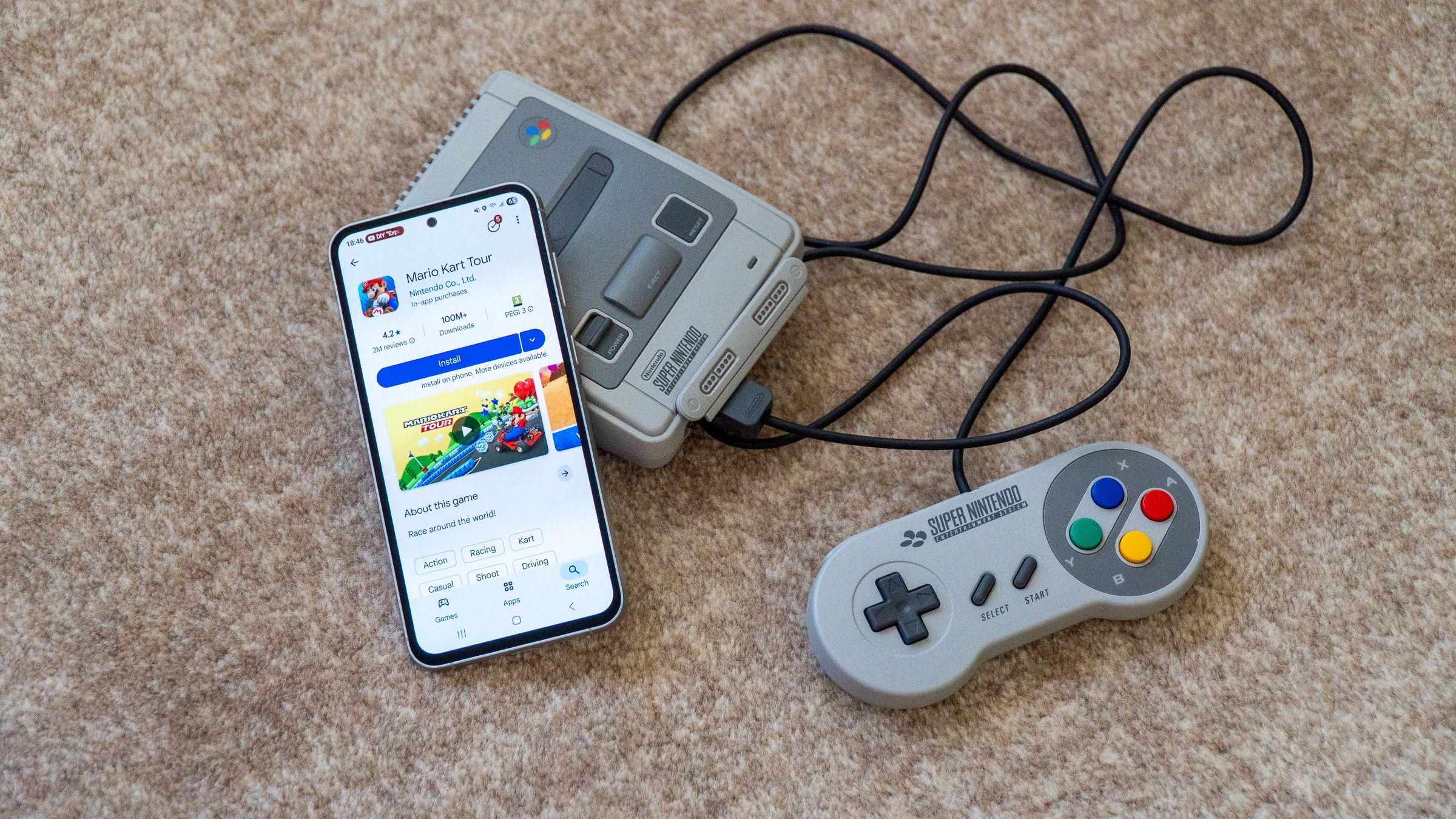 a phone displaying a mario game in the play store next to a classic super nintendo game console with a joystick on a carpet