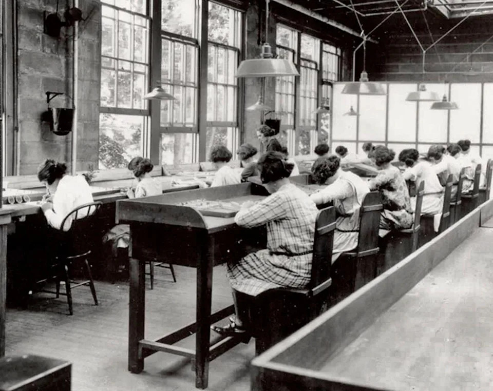 Women work at long tables in a historic factory setting, assembling small items, likely in the early 20th century