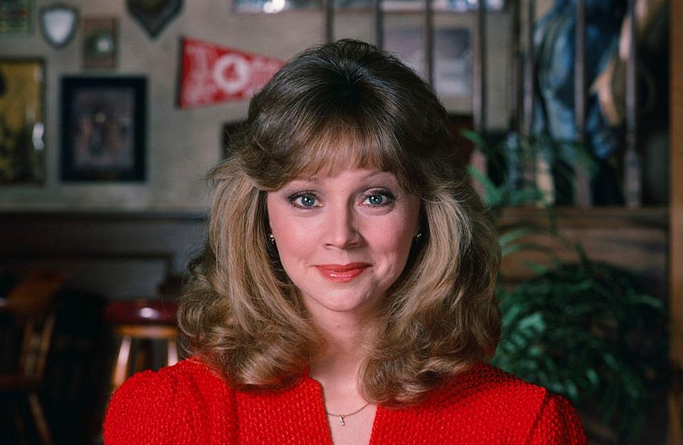Person with wavy hair smiling indoors, wearing a textured top; background shows various wall decorations
