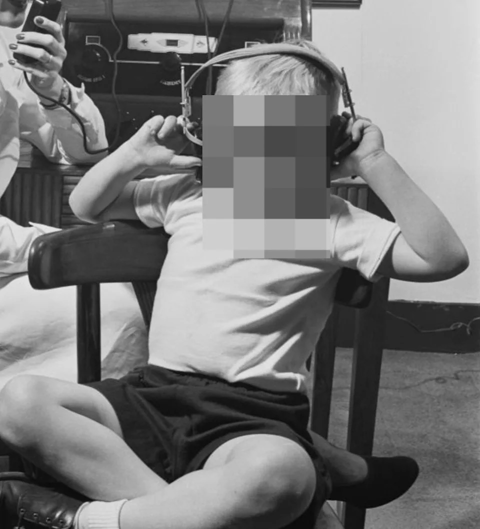 Woman in a lab coat uses a device to test young boy's hearing with headphones, seated in a clinical setting. Diagram of ear anatomy displayed behind