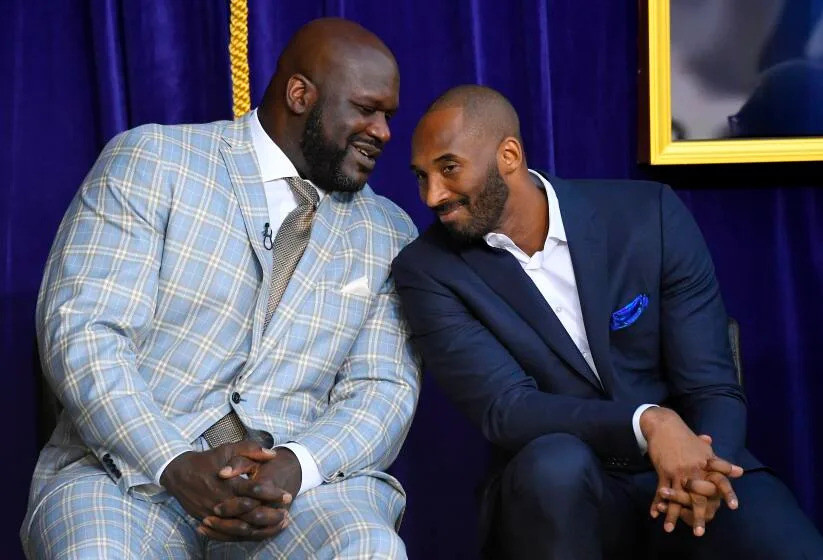 Kobe Bryant leans in to listen to Shaquille O'Neal as they sit onstage. They are both wearing suits, Kobe without a tie.