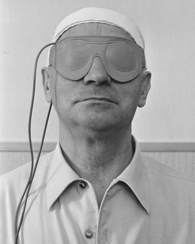 Person wearing a shirt and futuristic eye gear with wires attached, possibly engaging in a scientific or medical experiment